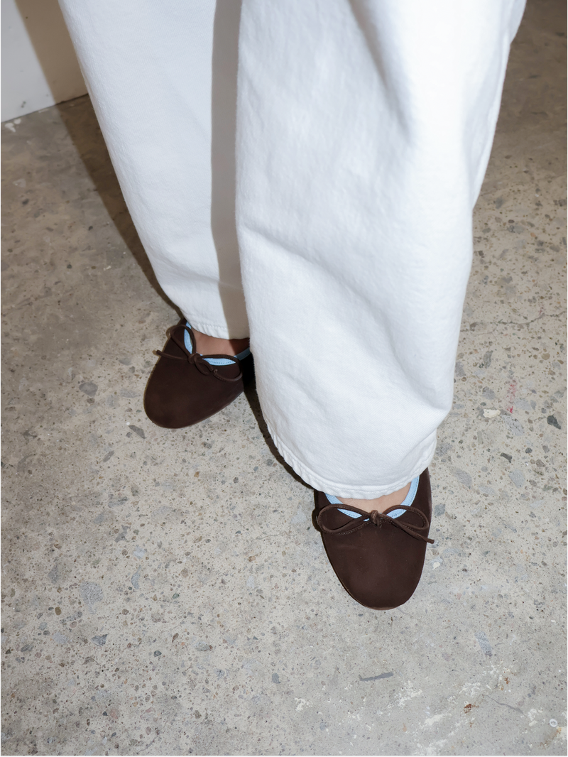 Brown loafers with blue accents worn with white pants on a concrete floor.