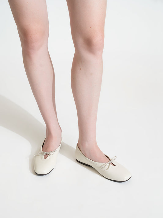 A person wearing a pair of off-white ballet flats with bow details, shown from the knees down against a clean white studio background.