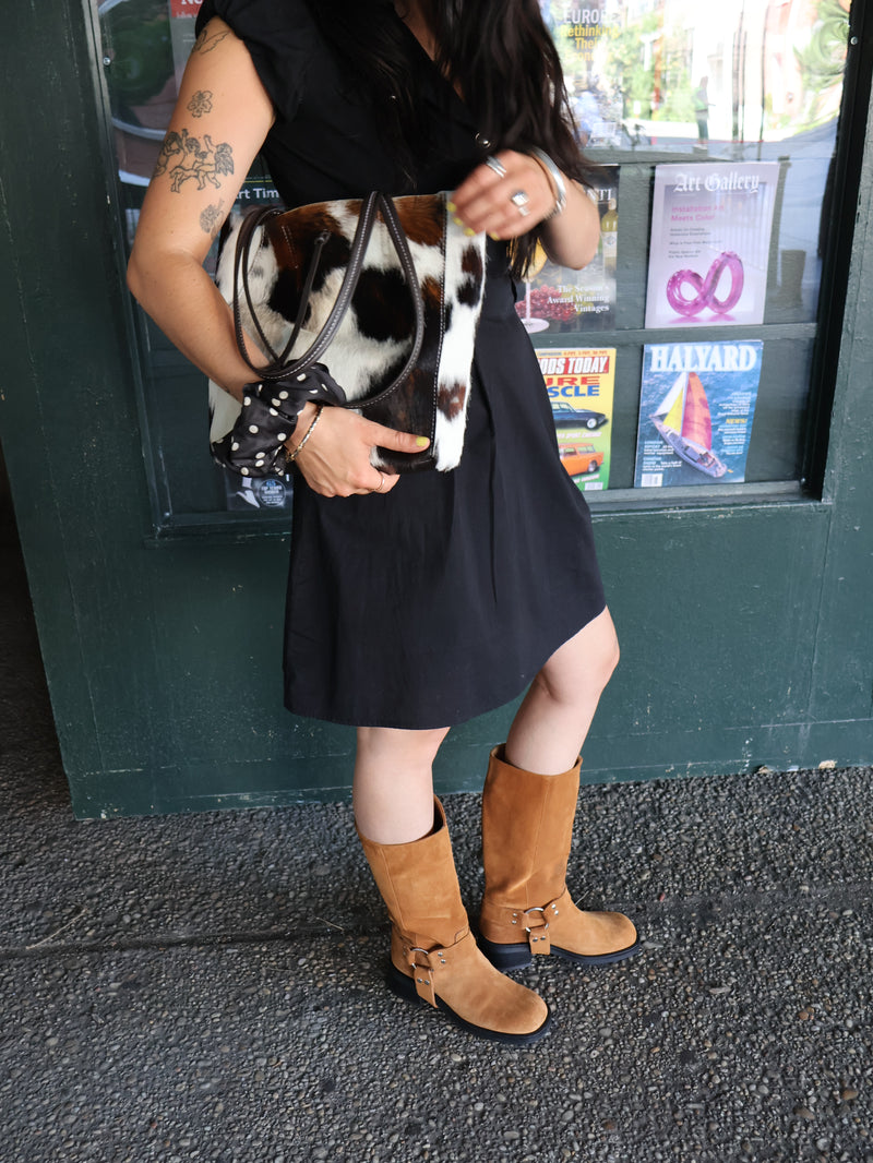 Mile-End fashionista wearing suede tan biker boots and a vintage black dress, while holding the cow-print bag of the year, propped up against a newstand wall
