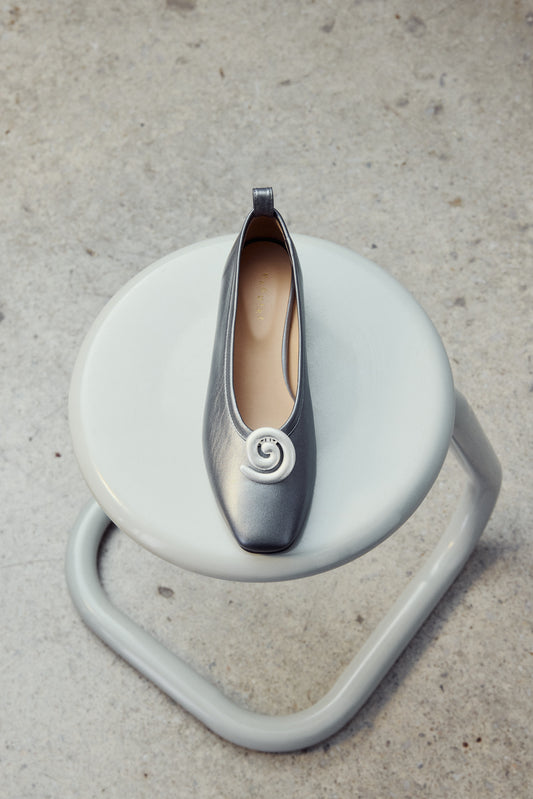Silver swirl shoe clip on grey ballet flat sitting on modern chair