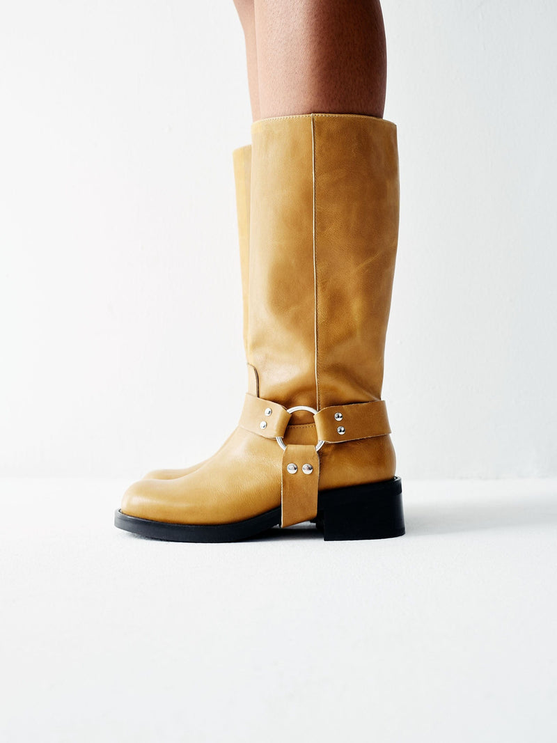 A yellow tan leather boot with a harness detail and low heel, displayed against a white background.