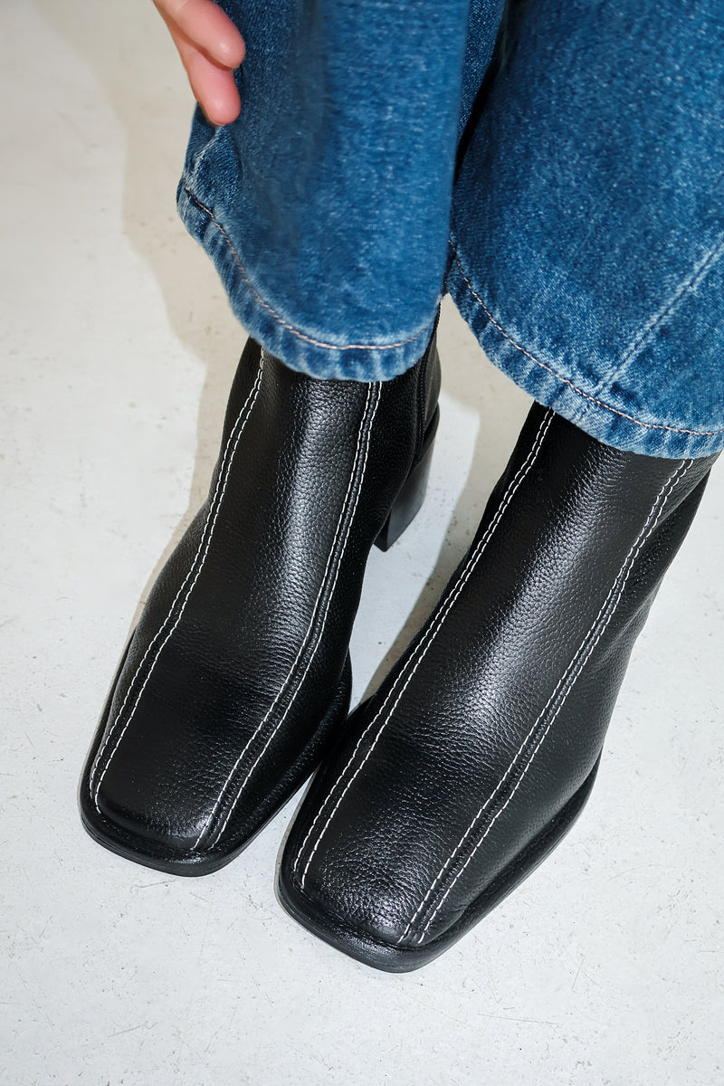 Close up imagine of black boots with white stitching, worn by woman wearing jeans on white floor